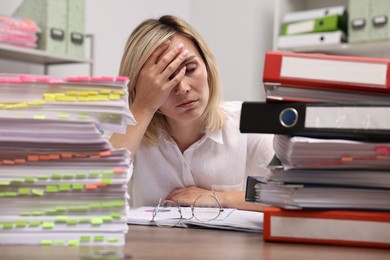 Overwhelmed woman sitting at table with stacks of documents and folders in office