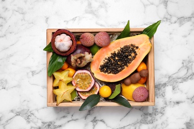 Photo of Different tropical fruits in wooden box on white marble table, top view