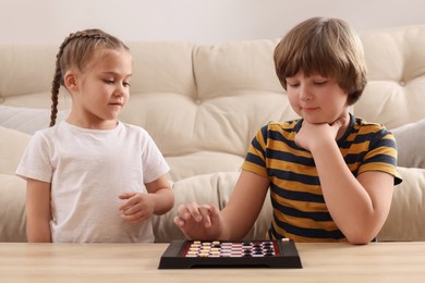 Photo of Cute boy playing checkers with little girl at light wooden table in room