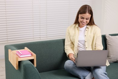 Happy woman using laptop on sofa with wooden armrest table at home