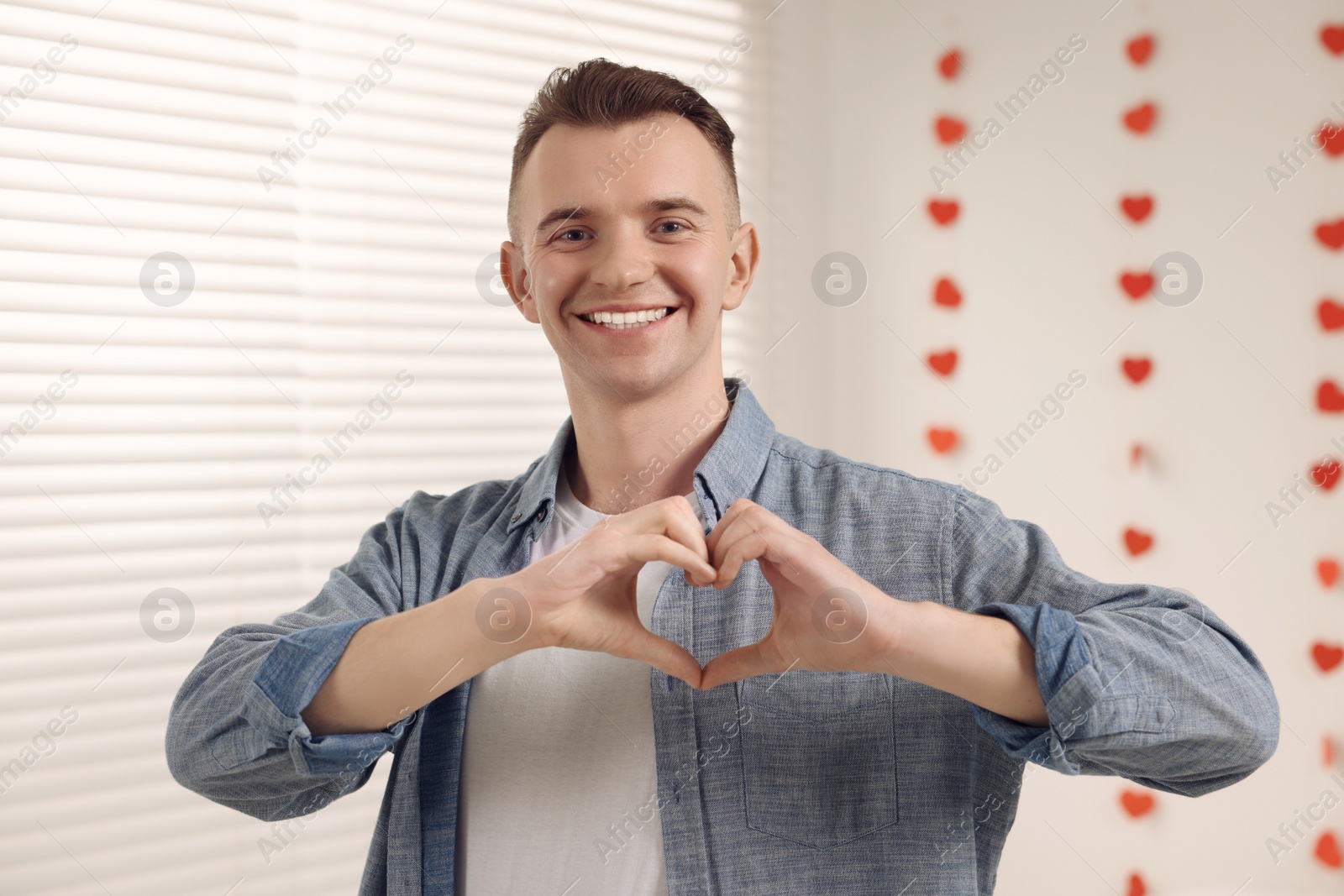 Photo of Handsome young man making heart with hands indoors, view from camera. Valentine's day celebration in long distance relationship