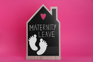 Photo of Wooden house figure with words Maternity Leave and paper cutout of baby feet on pink background