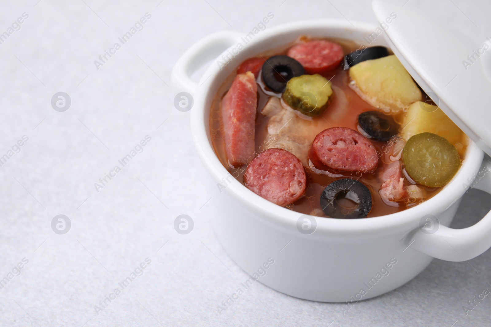 Photo of Meat solyanka soup with thin dry smoked sausages in pot on light table, closeup. Space for text