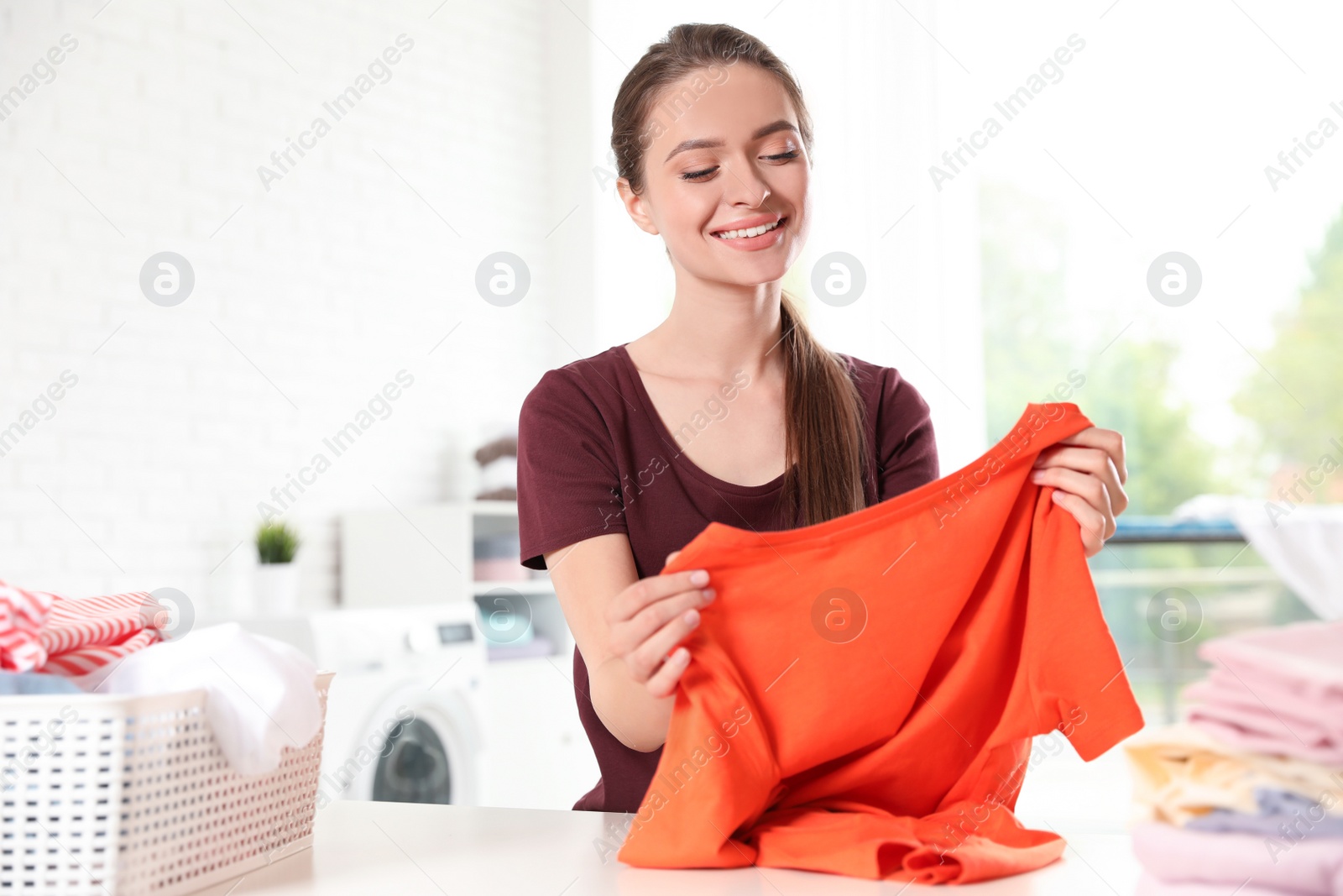 Photo of Happy young woman with clean t-shirt at table. Laundry day