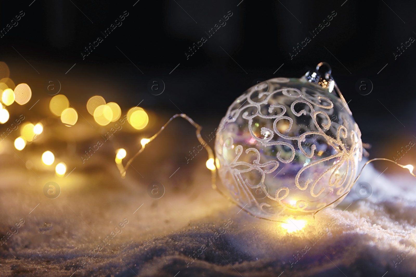 Photo of Christmas ball and fairy lights on white snow outdoors. Space for text