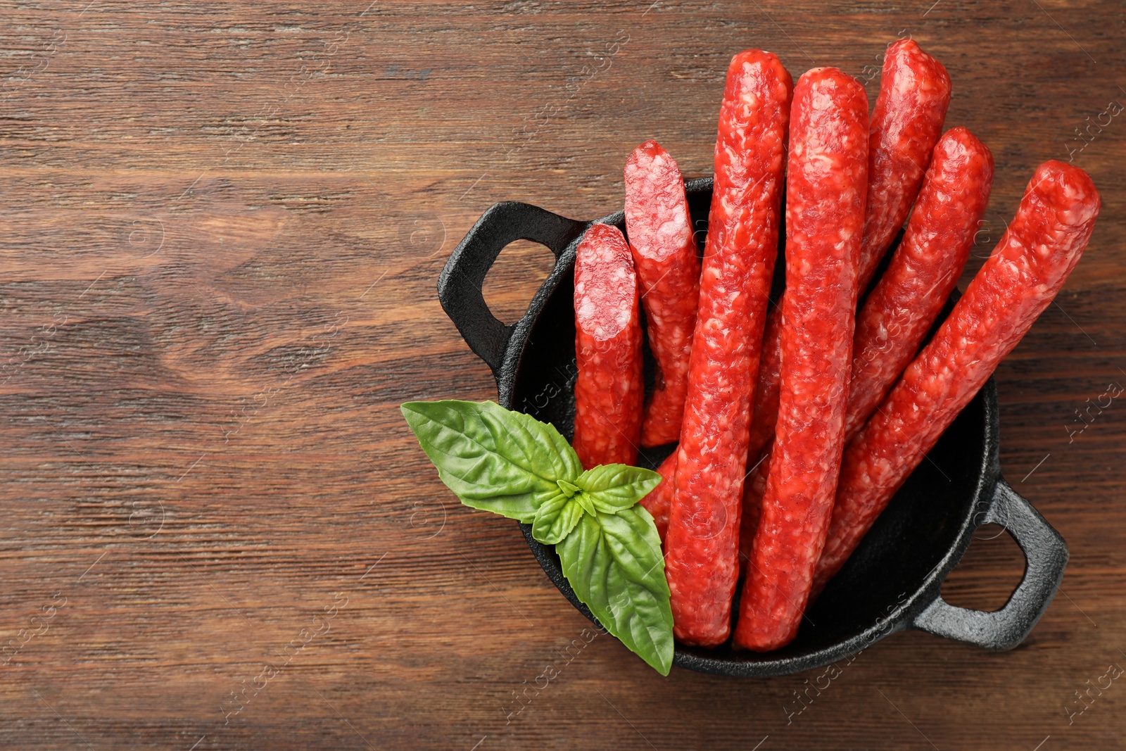 Photo of Thin dry smoked sausages and basil on wooden table, top view. Space for text