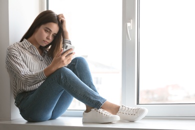 Photo of Young woman with mobile phone sitting on window sill, space for text. Loneliness concept