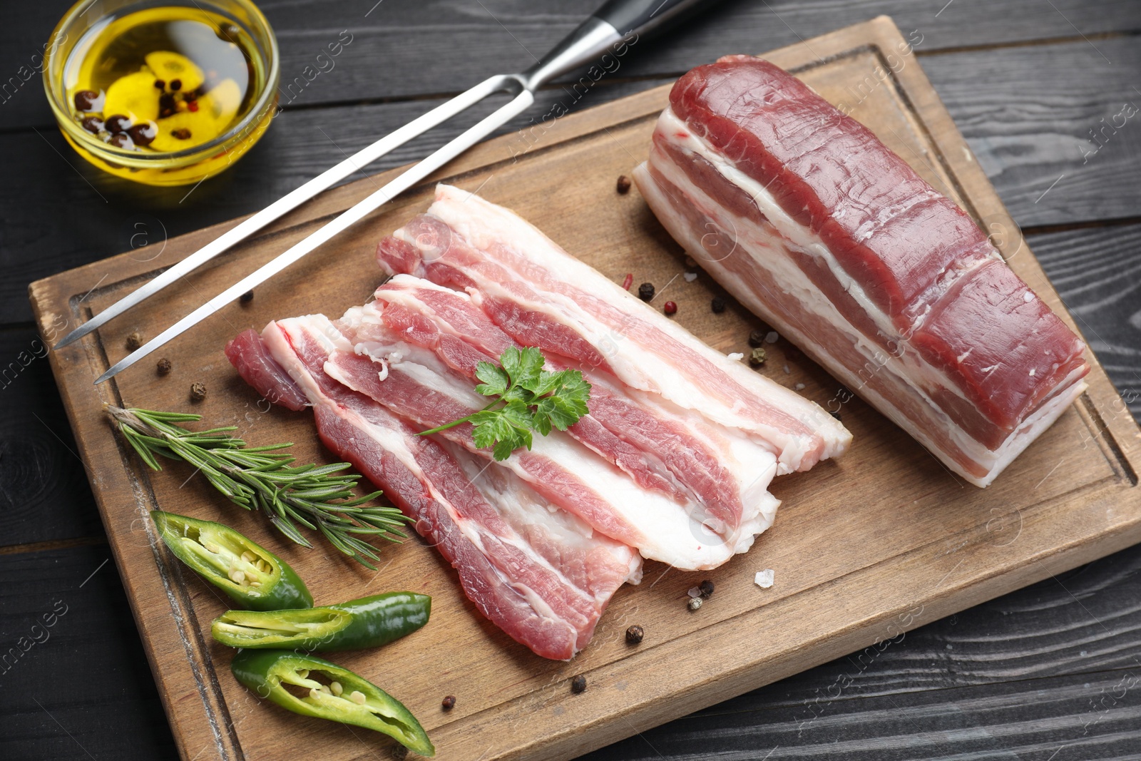 Photo of Pieces of raw pork belly, chili pepper, peppercorns, rosemary and parsley on black wooden table, above view