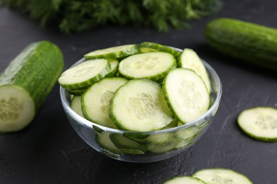 Photo of Cut cucumber in glass bowl and fresh vegetables on dark gray textured table, closeup
