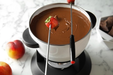 Dipping strawberry into pot with chocolate fondue on marble table