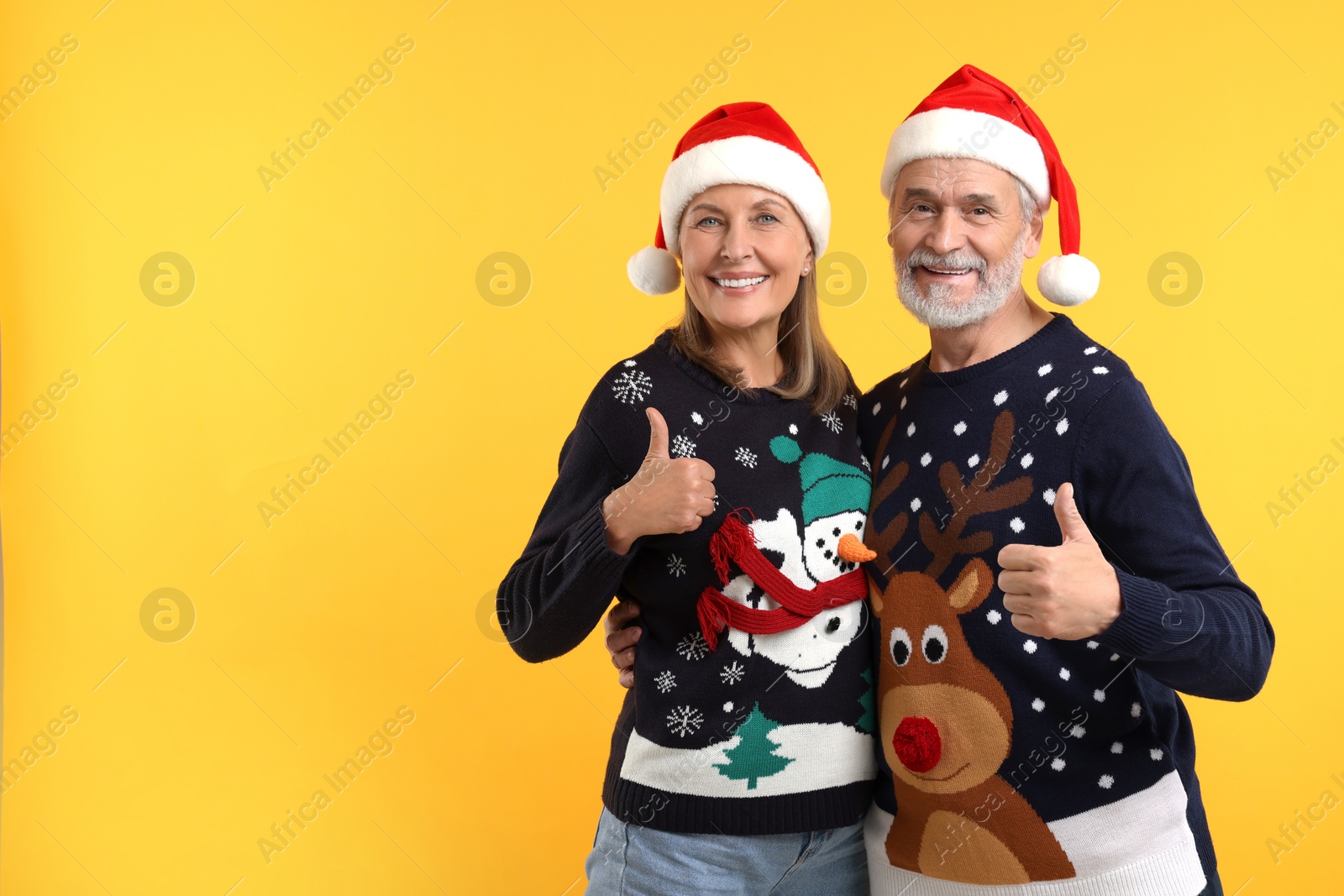 Photo of Happy senior couple in Christmas sweaters and Santa hats showing thumbs up on orange background. Space for text