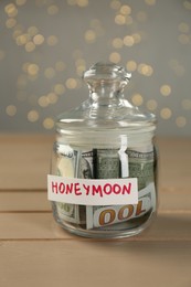 Glass jar with word Honeymoon and dollar banknotes on wooden table, closeup
