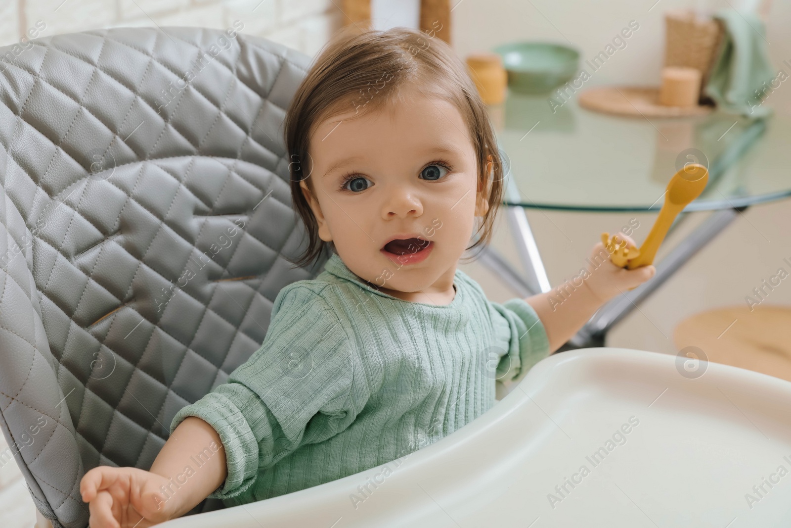 Photo of Cute little baby with cutlery sitting in high chair indoors