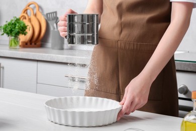 Woman sieving flour into baking dish at table in kitchen, closeup