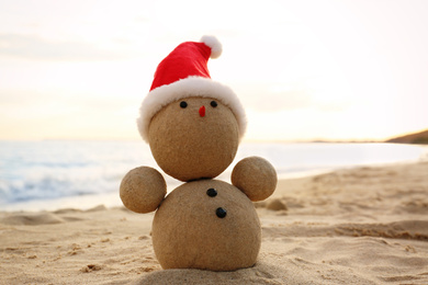Photo of Snowman made of sand with Santa hat on beach near sea at sunset. Christmas vacation