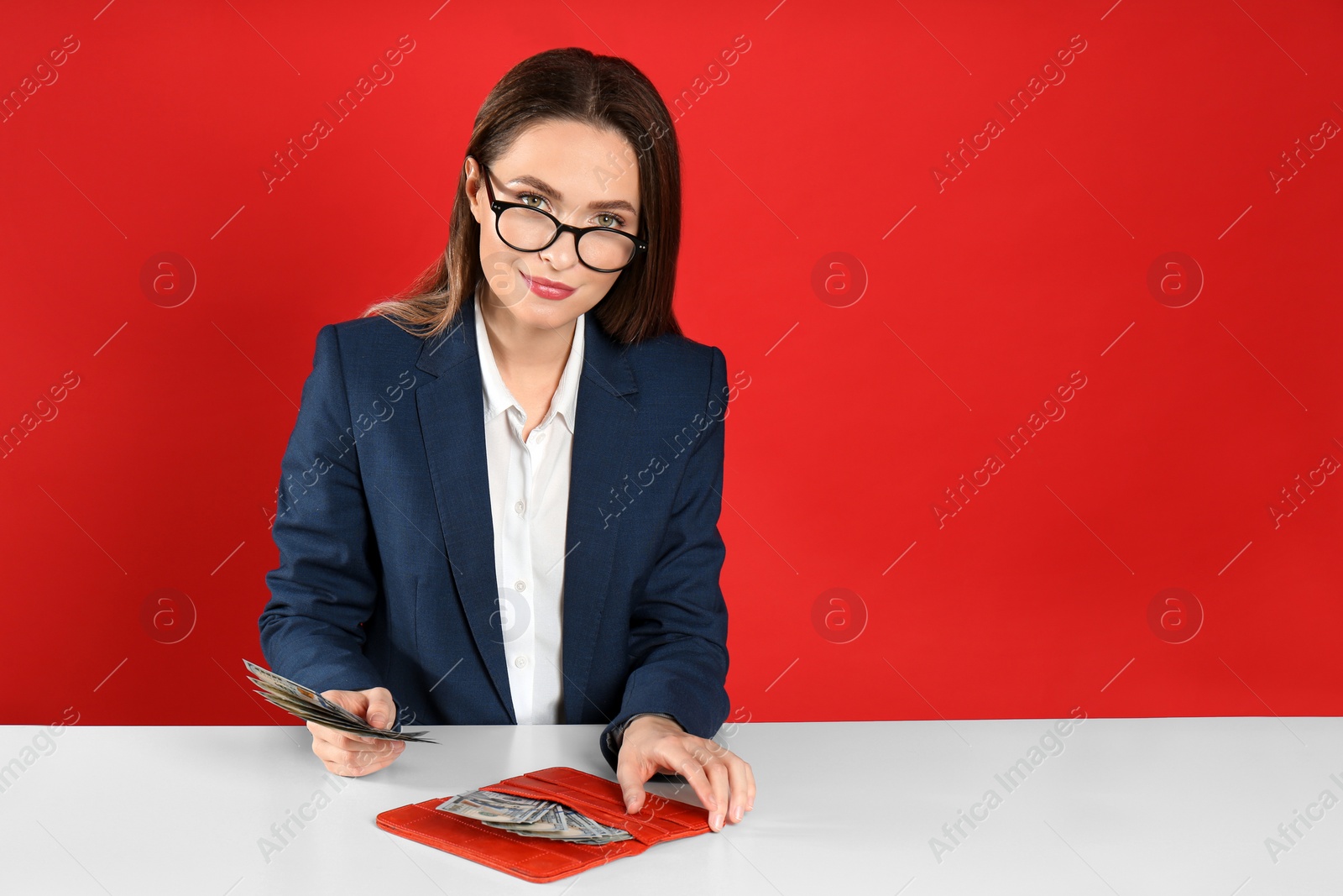 Photo of Young woman with money and wallet at table on crimson background. Space for text