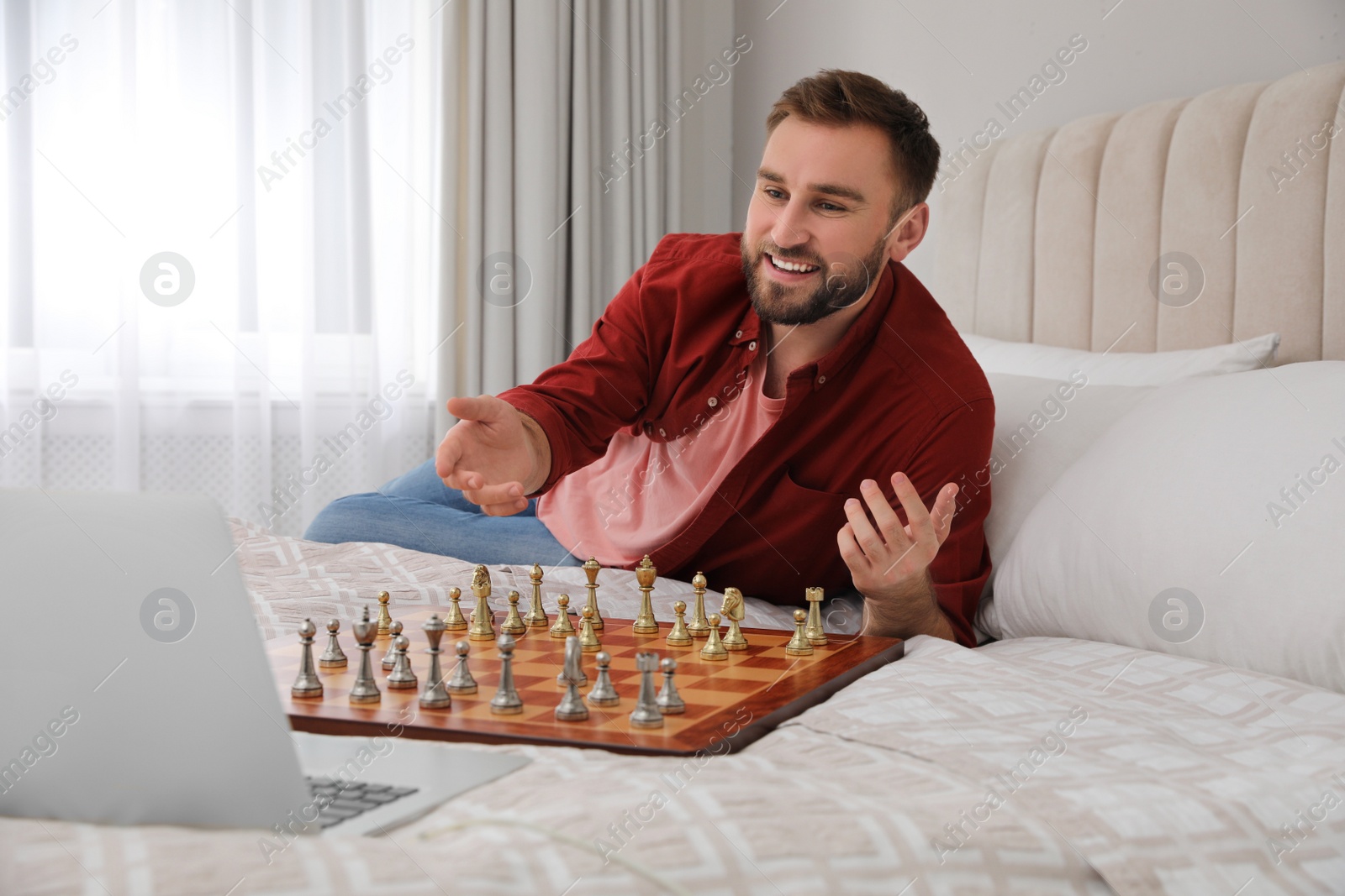 Photo of Young man playing chess with partner through online video chat on bed at home