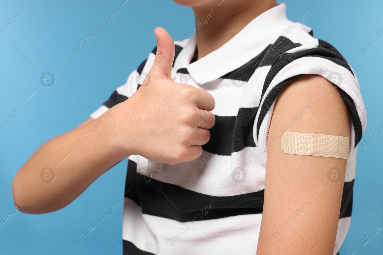 Photo of Boy with sticking plaster on arm after vaccination showing thumbs up against light blue background, closeup