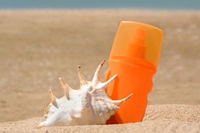 Bottle with sun protection spray and seashell on sandy beach, closeup
