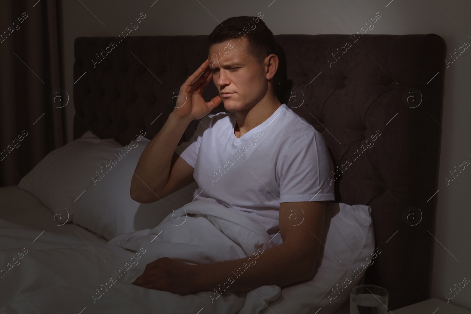 Photo of Handsome young man suffering from headache while sitting in bed at night