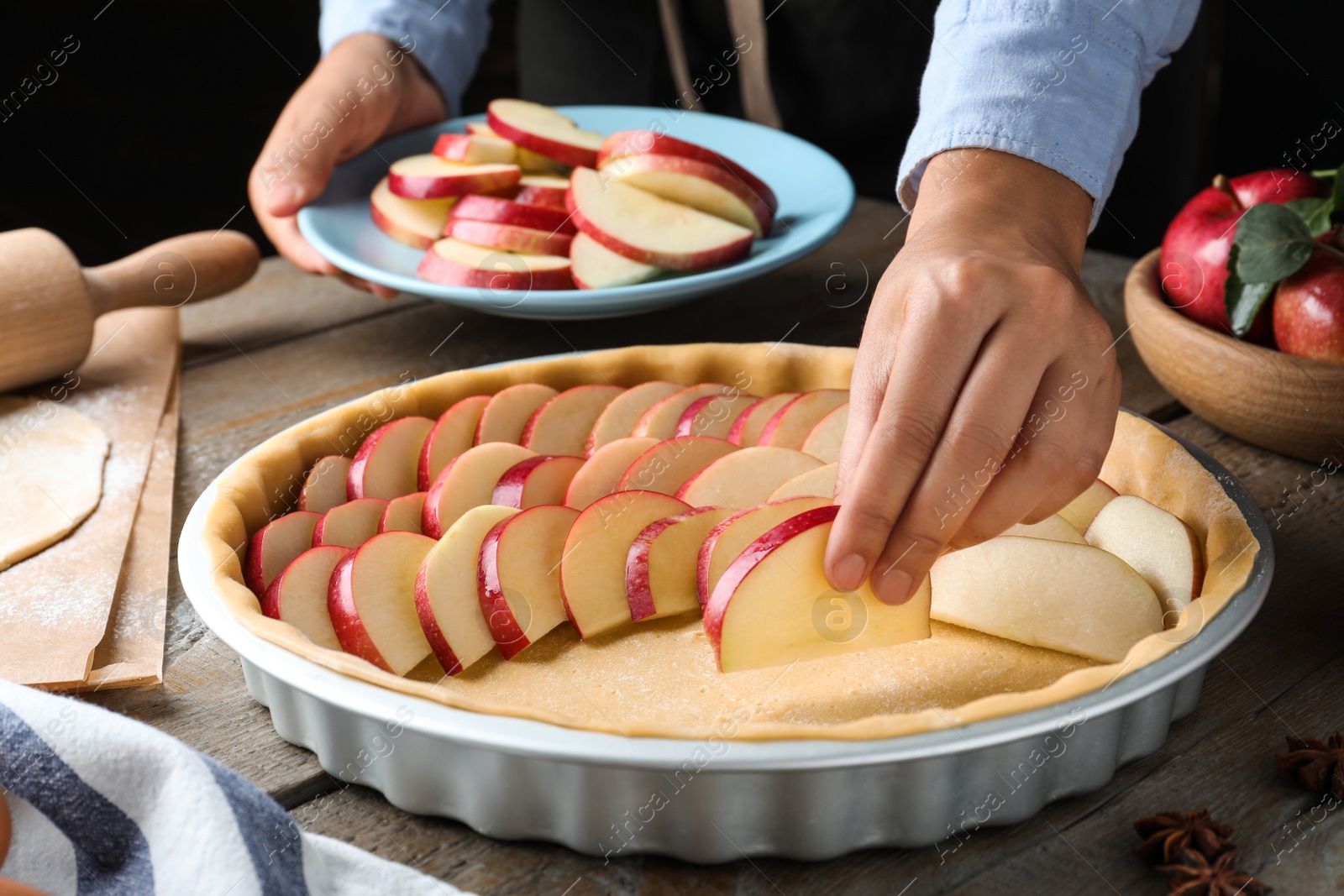 Photo of Woman putting apple slices into dish with raw dough at wooden table, closeup. Baking pie