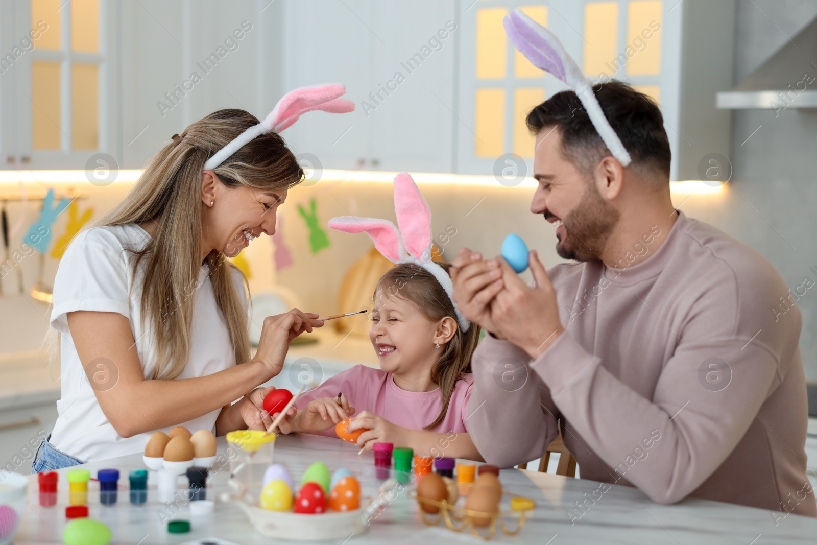 Photo of Easter celebration. Happy family with bunny ears having fun while painting eggs at white marble table in kitchen