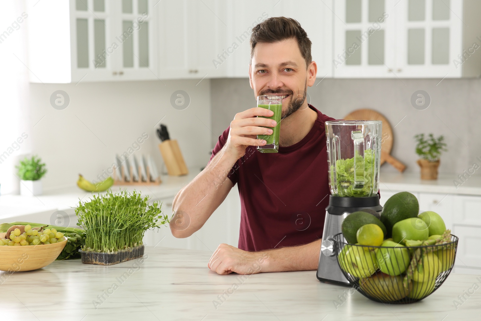 Photo of Happy man drinking delicious smoothie at white marble table in kitchen