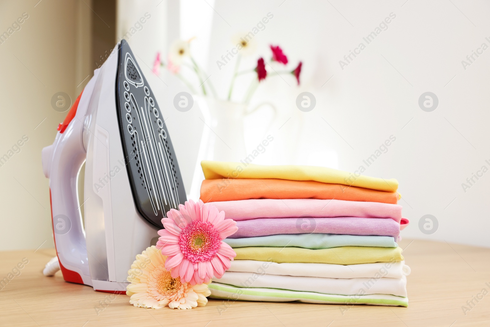 Photo of Stack of clean clothes, modern iron and flowers on wooden table