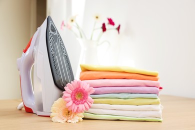 Photo of Stack of clean clothes, modern iron and flowers on wooden table