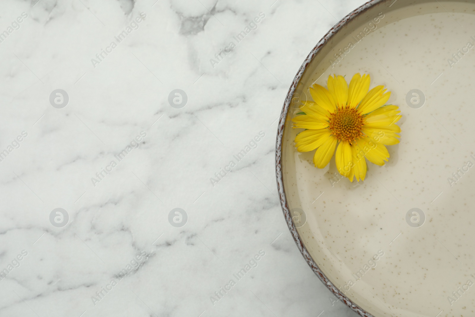 Photo of Beige bowl with water and yellow flower on white marble table, top view. Space for text