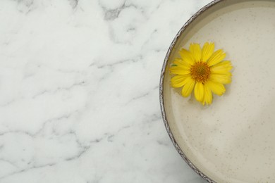 Photo of Beige bowl with water and yellow flower on white marble table, top view. Space for text