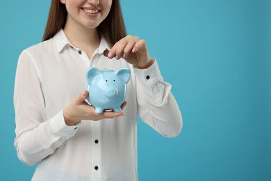 Woman putting coin into piggy bank on light blue background, closeup. Space for text