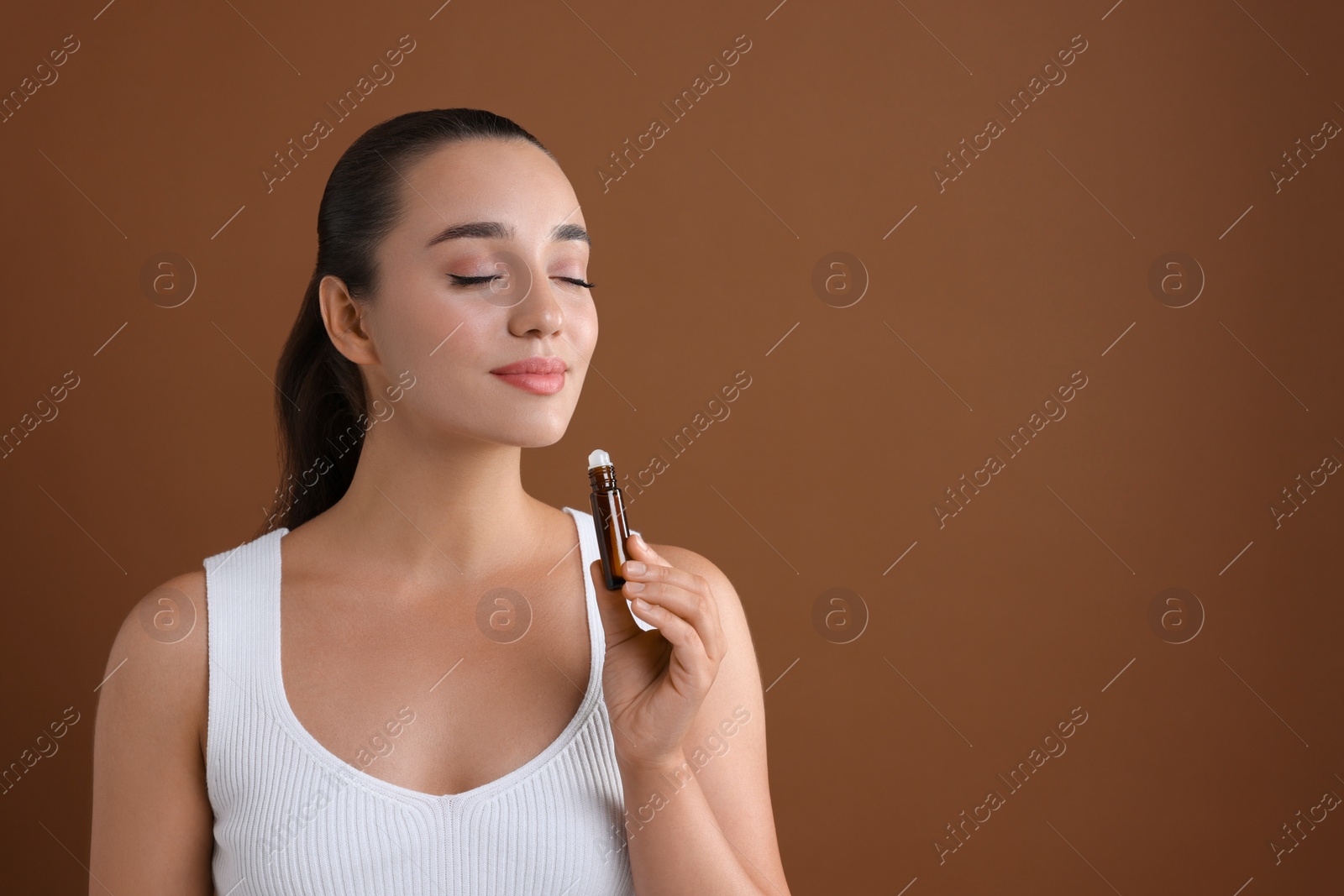 Photo of Beautiful young woman with roller bottle of essential oil on brown background, space for text