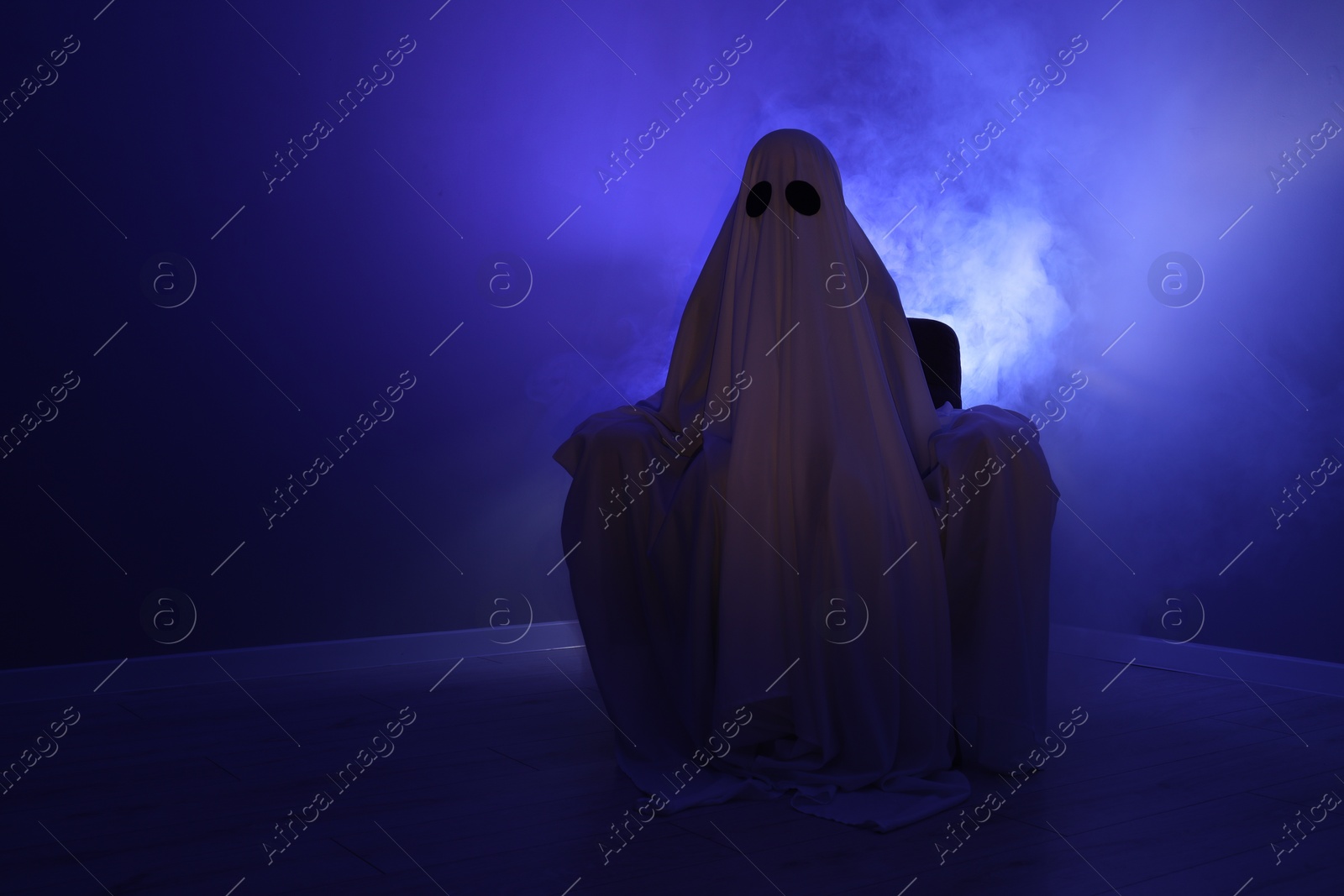 Photo of Creepy ghost. Woman covered with sheet sitting in armchair in blue light, space for text