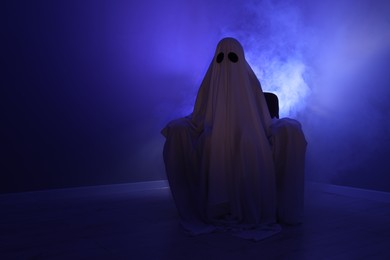 Photo of Creepy ghost. Woman covered with sheet sitting in armchair in blue light, space for text