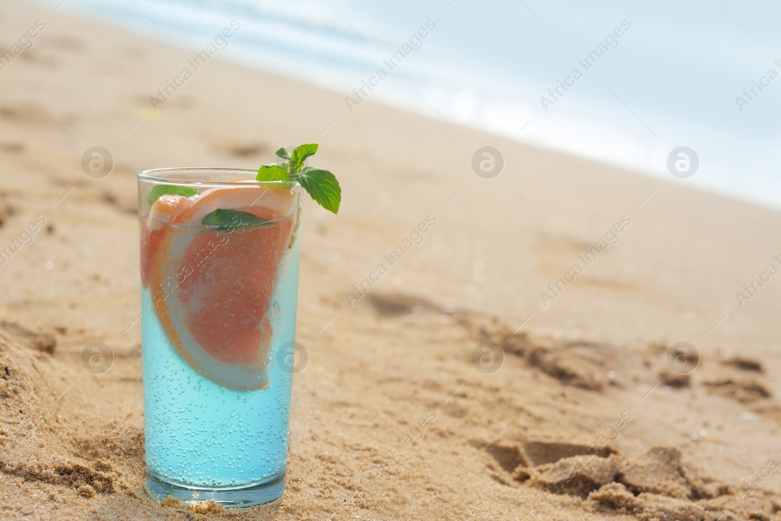 Photo of Glass of refreshing drink with grapefruit and mint on sandy beach near sea, space for text
