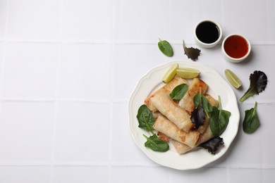 Photo of Plate with tasty fried spring rolls, spinach, lime and sauces on white tiled table, flat lay. Space for text