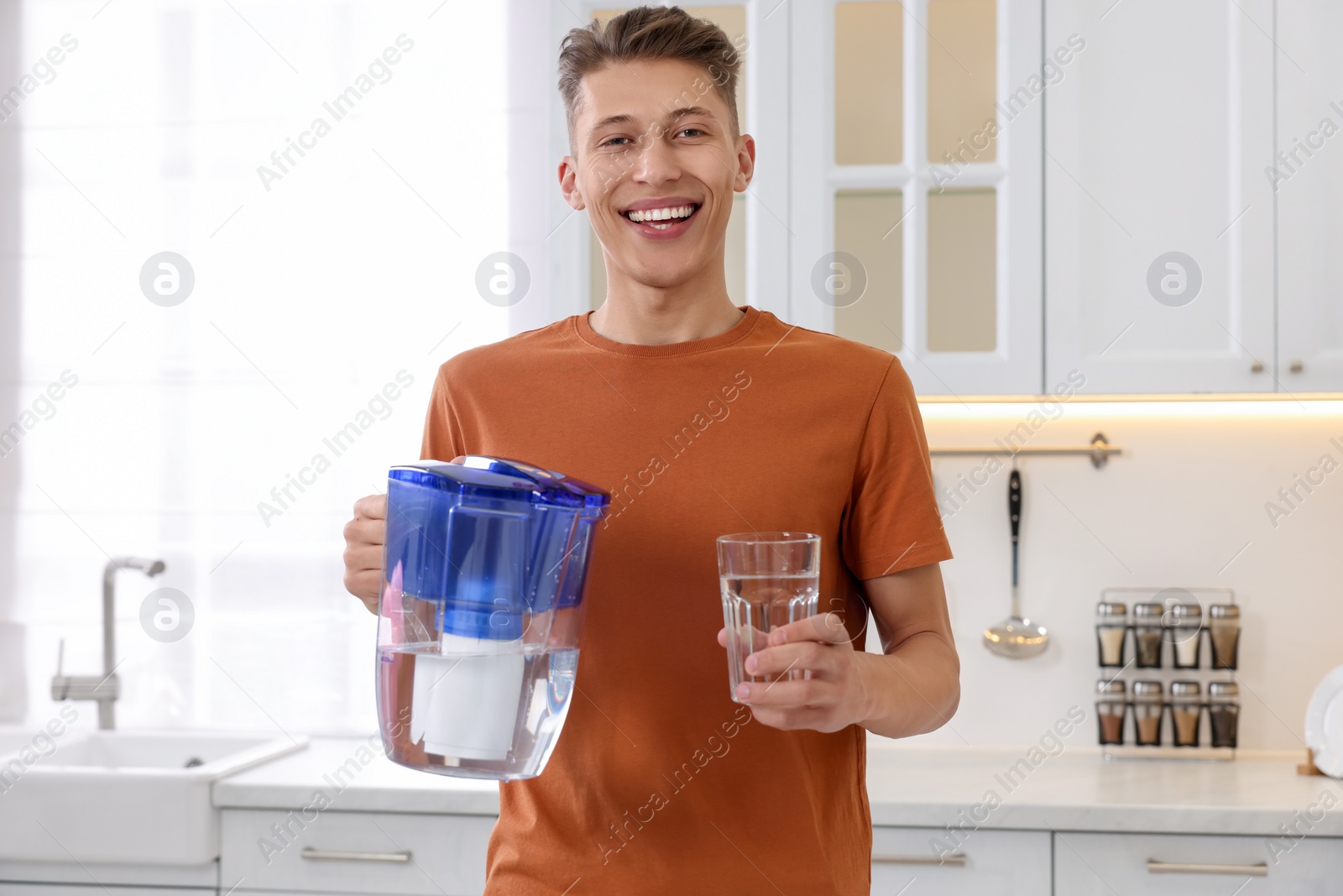 Photo of Happy man with filter jug and glass of clear water in kitchen