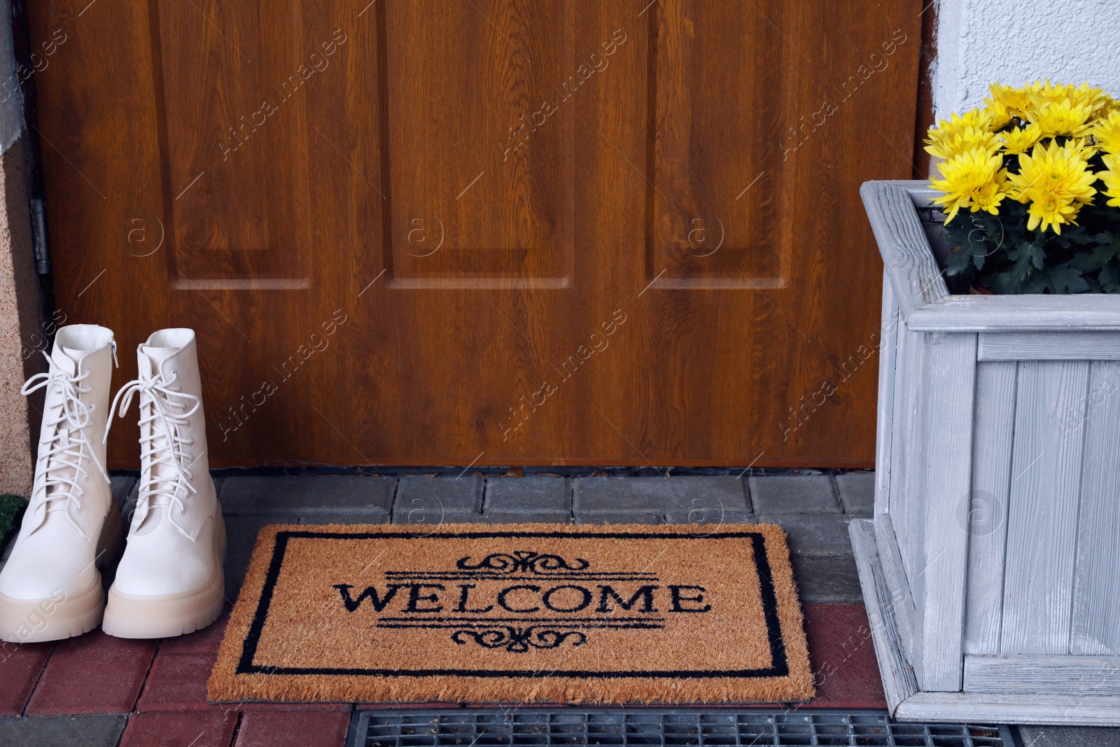 Photo of Doormat with word Welcome, stylish boots and beautiful flowers on floor near entrance