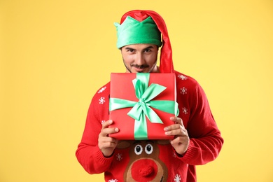 Photo of Happy man in Christmas sweater and elf hat holding gift box on yellow background