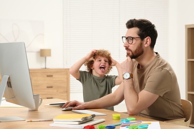 Photo of Little boy bothering his father at home. Man working remotely at desk