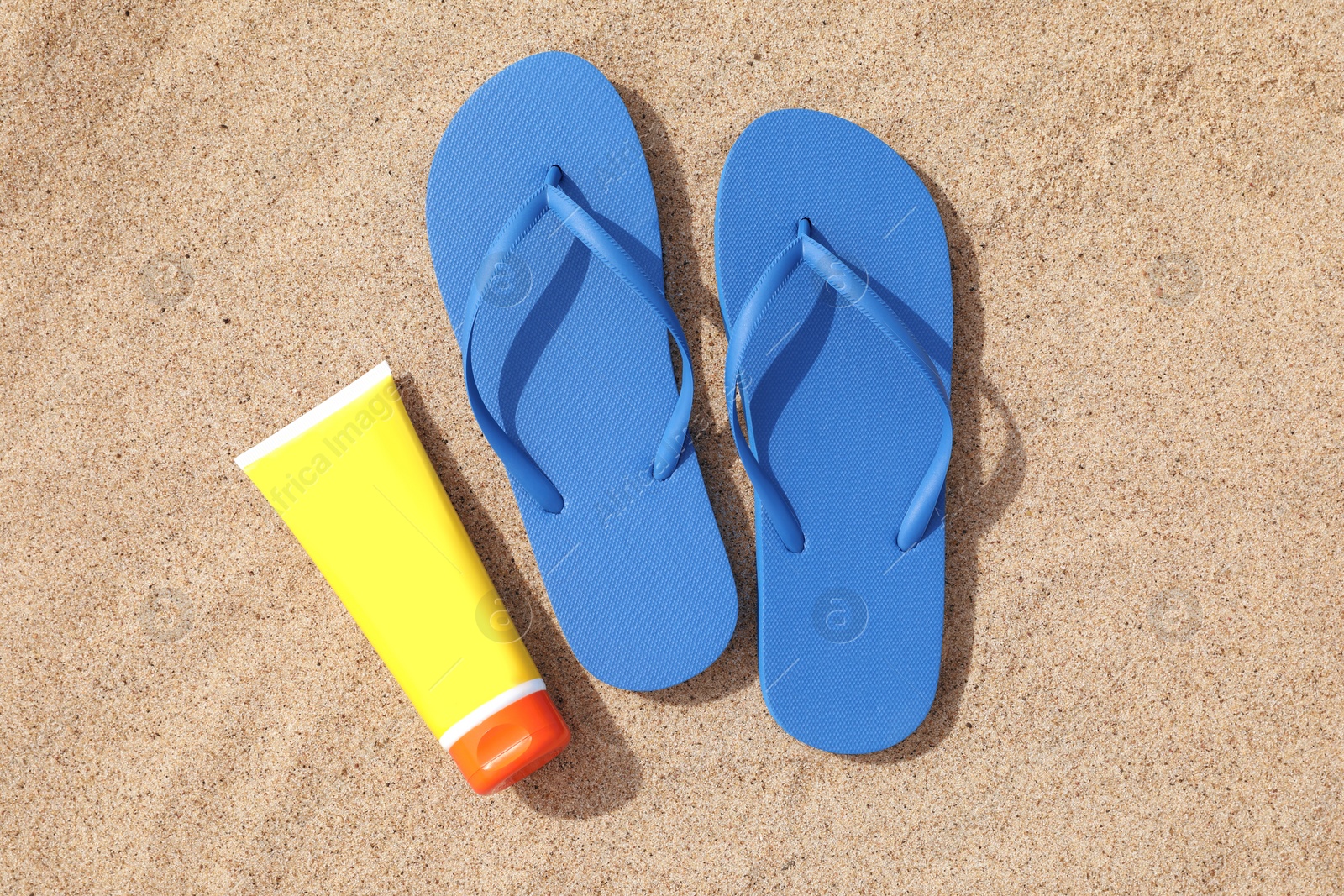 Photo of Blue flip flops and sunscreen on sand, flat lay