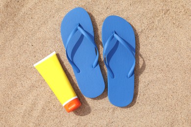 Blue flip flops and sunscreen on sand, flat lay