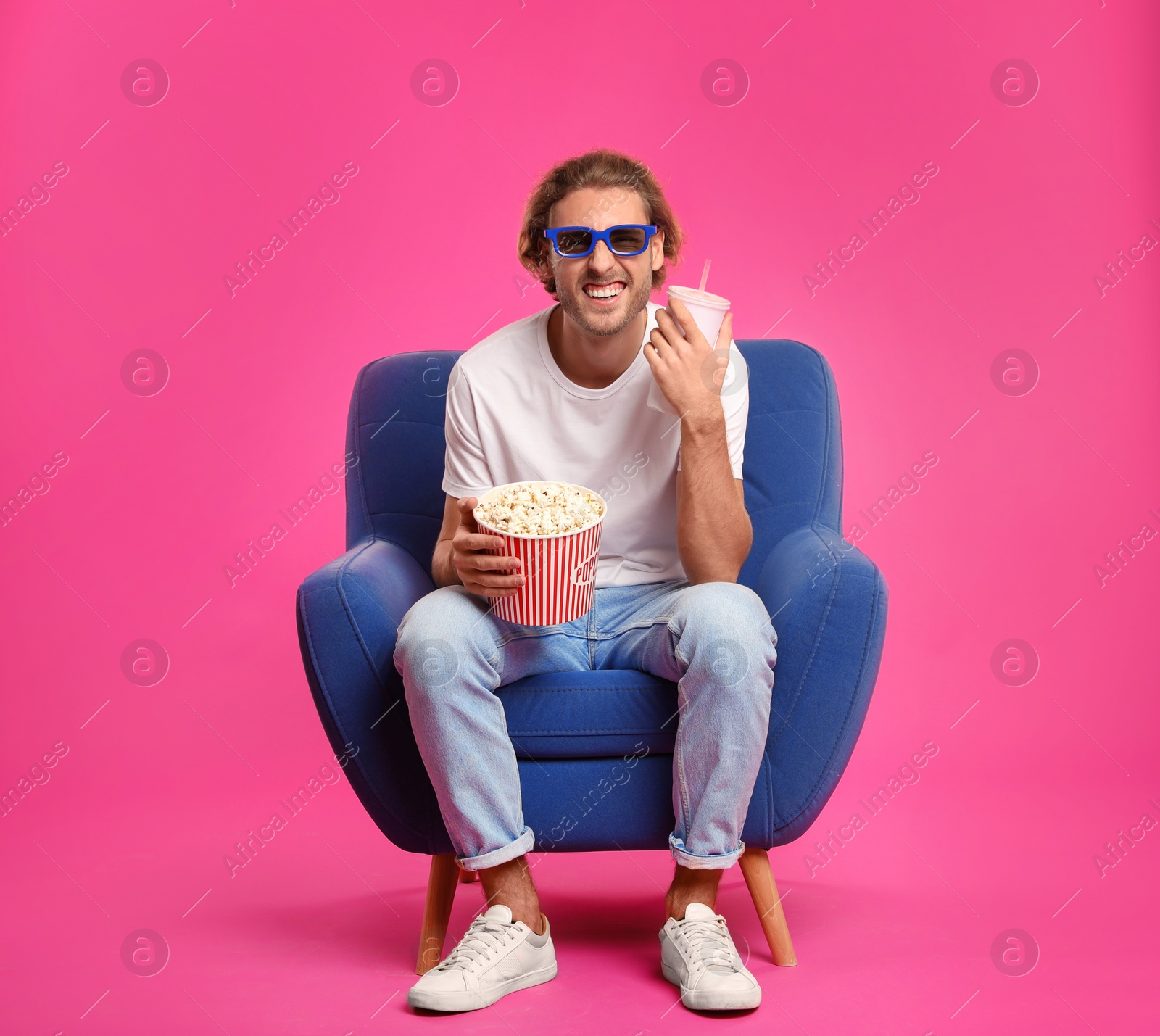 Photo of Man with 3D glasses, popcorn and beverage sitting in armchair during cinema show on color background