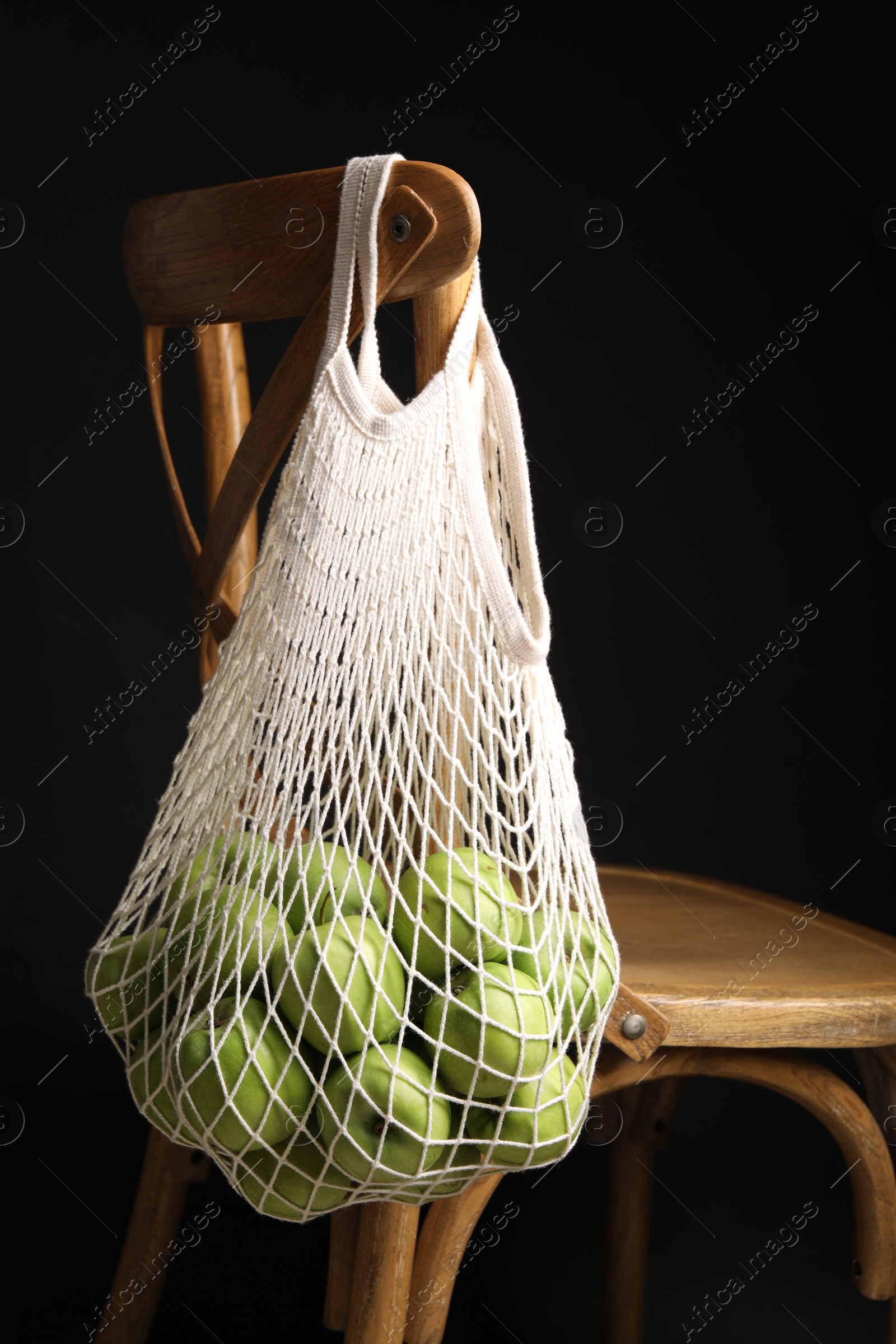 Photo of Green apples in net bag hanging on wooden chair against black background