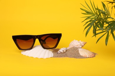 Stylish sunglasses, seashells, sand and palm leaves on yellow background