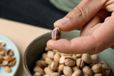 Woman holding tasty peeled pistachio nut over bowl, closeup