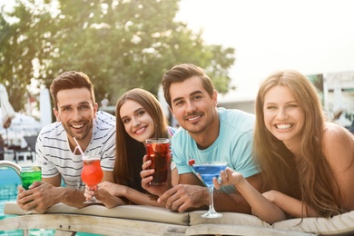 Happy young friends with fresh summer cocktails relaxing near swimming pool