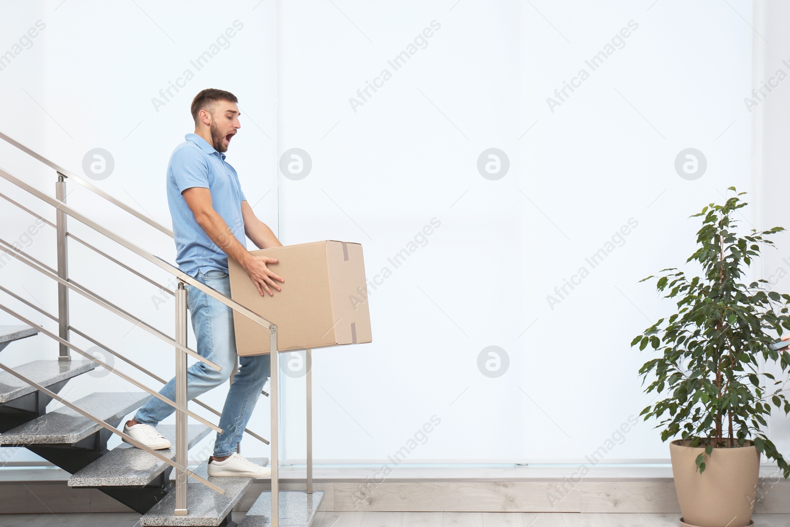 Photo of Clumsy young man carrying carton box downstairs indoors. Posture concept