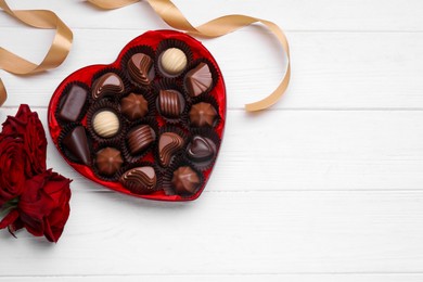 Heart shaped box with delicious chocolate candies, roses and ribbon on white wooden table, flat lay. Space for text